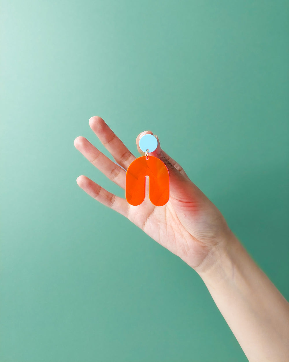 Hand hält leichten orangefarbenen Acryl Ohrring in Bogenform mit Ohrstecker vor grünem Hintergrund